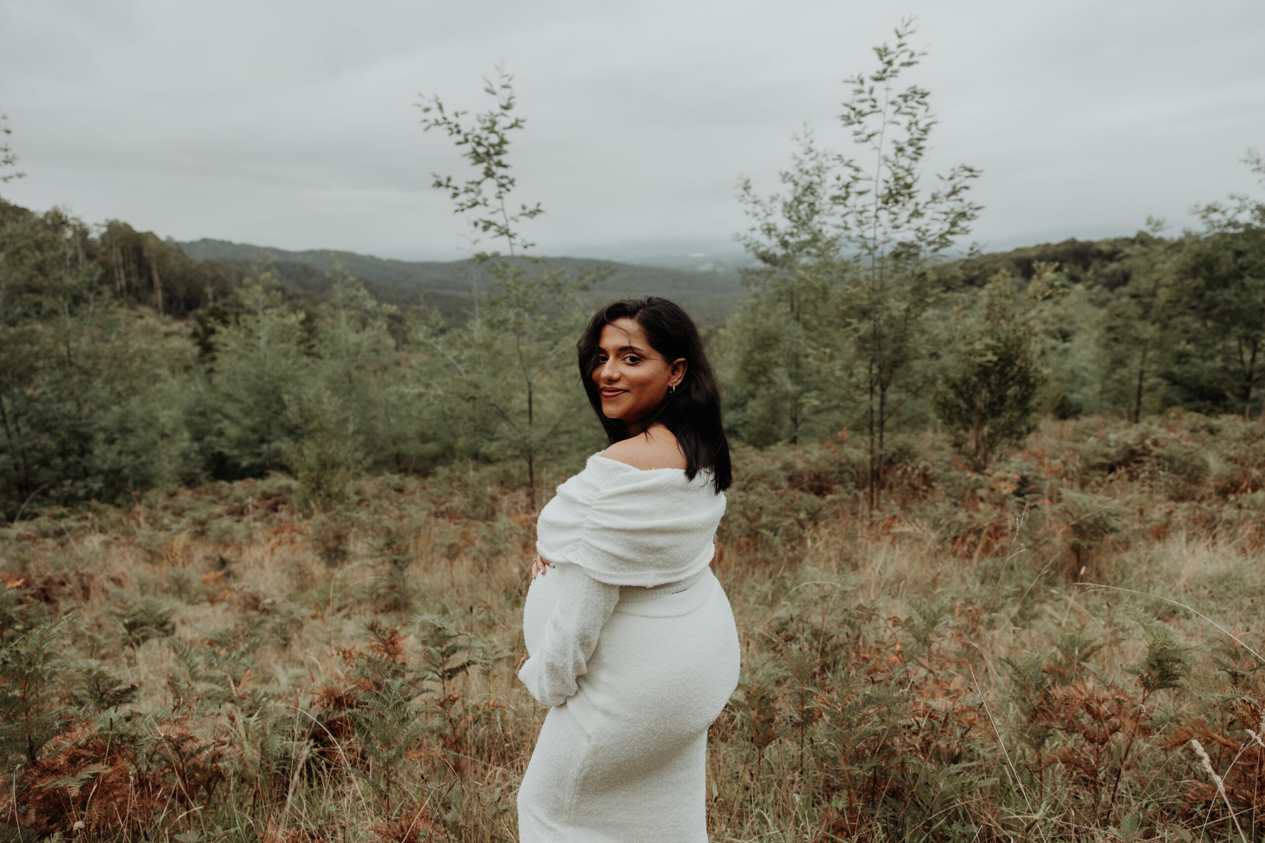 Maternity photography session held in Melbourne Dandenong Ranges captures pregnant woman in white knit dress standing against hill backdrop